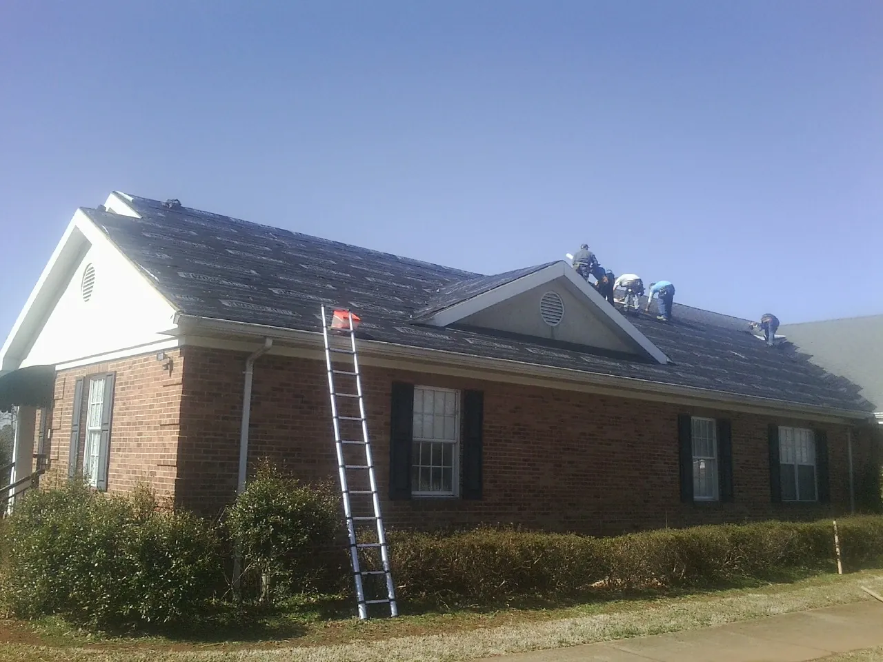 Crew tearing shingles off a brick ranch roof with a ladder set — Metal Roof Repair work by our crew in Calhan