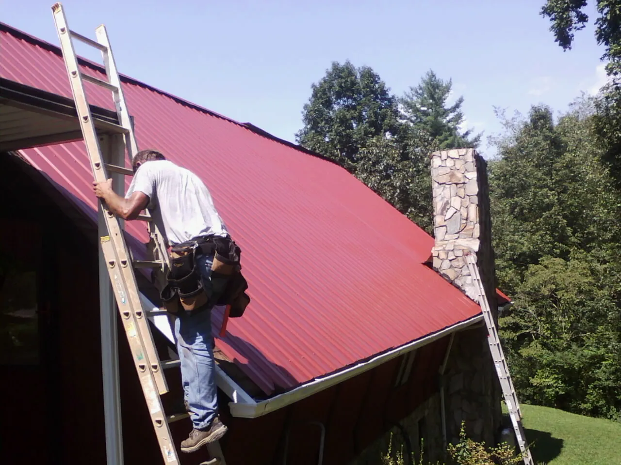 Roofer on a ladder beside a red metal roof and stone chimney — Soffit & Fascia Repair by Roofing Brothers in Calhan